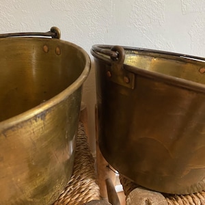 May include: Two round brass buckets with dark metal swing handles attached by riveted brackets. The buckets feature a polished metallic finish with visible signs of wear and patina, showing a smooth, curved construction and open tops.