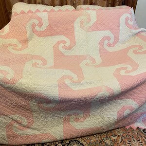 May include: A large quilt featuring a repeating geometric pattern of interlocking spiral shapes in soft pink and off-white fabric. The entire surface is covered in a fine, diamond-shaped grid of stitching, creating a textured appearance.