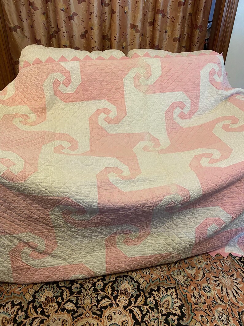 May include: A large quilt featuring a repeating geometric pattern of interlocking spiral shapes in soft pink and off-white fabric. The entire surface is covered in a fine, diamond-shaped grid of stitching, creating a textured appearance.