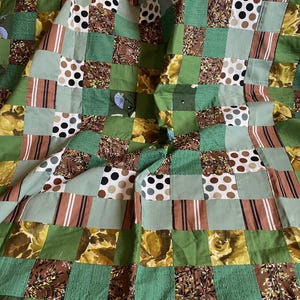 May include: A patchwork quilt featuring a variety of green, brown, and cream-colored fabrics. The quilt is composed of square blocks with patterns including polka dots, floral prints, and stripes. The overall design is a mix of textures and shades of green.