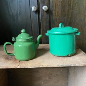Könnte beinhalten: Eine grüne Emaille-Teekanne und ein kleiner Topf mit Deckel stehen auf einer Holzoberfläche. Die Teekanne hat einen gebogenen Ausguss und einen Griff. Der Topf hat zwei seitliche Griffe und einen Griff am Deckel. Beide Artikel sind in leuchtendem Grün lackiert.