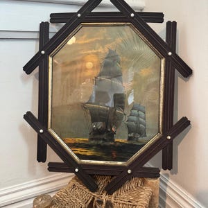 Może przedstawiać: Antyczny, oprawiony wydruk marynistyczny przedstawiający dwa żaglowce na oceanie o zachodzie słońca. Ciemnobrązowa rama ma kształt ośmiokąta z dekoracyjnymi akcentami w kształcie krzyża i małymi białymi perłami. Około 50 cm.