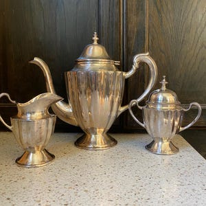 以下が含まれることがあります： 銀色のティーセット。ティーポット、クリーマー、砂糖入れが含まれています。ティーポットは湾曲した注ぎ口と装飾的なつまみ付きの蓋が付いています。クリーマーと砂糖入れには取っ手があり、まだらのカウンタートップに置かれています。