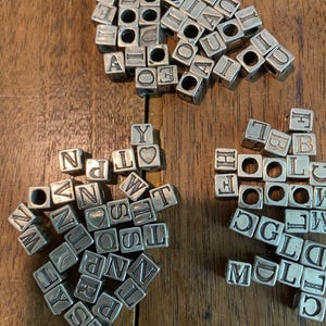 May include: Silver-toned metal alphabet beads with letters A-Z, in a variety of fonts. The beads are cube-shaped with a hole through the center, perfect for jewelry making or crafting. The beads are scattered on a wooden surface.