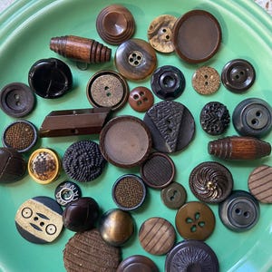 Puede incluir: Una colección de botones vintage en varios colores, formas y tamaños. Los botones son principalmente marrones, negros y dorados, y algunos tienen diseños intrincados.