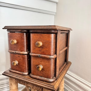 Puede incluir: Un pequeño mueble antiguo de madera con cuatro cajones, cada uno con un pomo redondo de color claro. El mueble es de un color marrón cálido y rico y se apoya sobre un soporte de estilo bambú. La veta de la madera es visible, lo que añade encanto vintage.