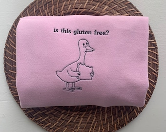 May include: A pink sweatshirt with a cartoon duck holding a slice of bread and the text "is this gluten free?" embroidered on the front.