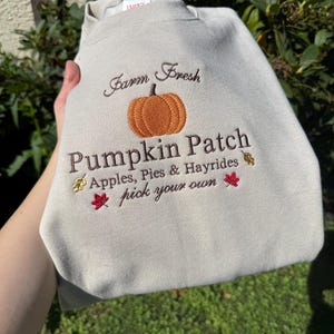 May include: A light beige crewneck sweatshirt featuring embroidered text that reads Farm Fresh Pumpkin Patch, Apples, Pies & Hayrides, and pick your own. The design includes a central orange pumpkin and small red and yellow leaf accents.