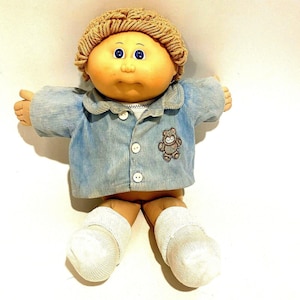 May include: A vintage Cabbage Patch Kid doll with a tan face, blue eyes, and light brown yarn hair. The doll is dressed in a blue corduroy jacket with a teddy bear patch and white socks.