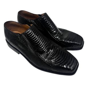 May include: A pair of black slip-on shoes with a textured, reptile-like pattern. The shoes have a square toe and a low heel. The leather appears shiny. The shoes are set against a white background.