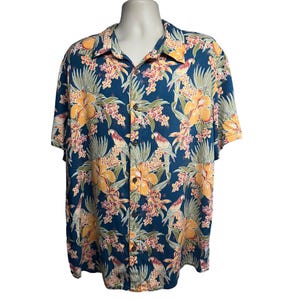 Puede incluir: Una camisa azul de manga corta con botones y estampado tropical con flores amarillas, hojas verdes y loros rosas y azules.