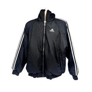 May include: Black Adidas jacket with white stripes on the sleeves. The jacket features a high collar, a zip closure, and the Adidas logo on the chest. It has a ribbed hem and cuffs.