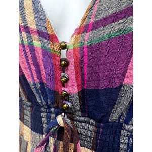 Pode incluir: Um vestido xadrez colorido com decote em V e bot&otilde;es cor de lat&atilde;o. O vestido apresenta uma cintura com cord&atilde;o e uma mistura de tons rosa, roxo, azul e bege. O tecido tem uma apar&ecirc;ncia texturizada.