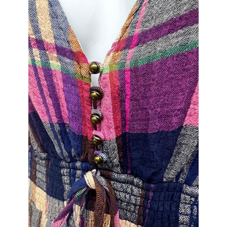 Pode incluir: Um vestido xadrez colorido com decote em V e bot&otilde;es cor de lat&atilde;o. O vestido apresenta uma cintura com cord&atilde;o e uma mistura de tons rosa, roxo, azul e bege. O tecido tem uma apar&ecirc;ncia texturizada.