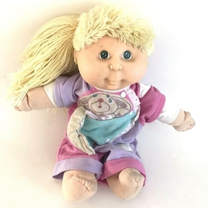 Könnte beinhalten: Eine Vintage Cabbage Patch Kids Puppe mit blonden Garnhaaren in einem Pferdeschwanz. Die Puppe trägt ein rosa, lila und türkisfarbenes Outfit mit einem Bärenmuster. Die Puppe hat grüne Augen und einen weichen Stoffkörper.