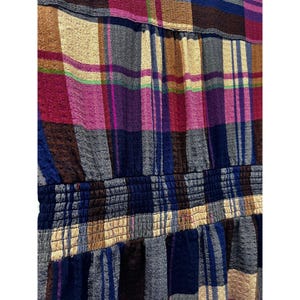 Pode incluir: Close-up de um tecido xadrez colorido. O padr&atilde;o apresenta uma mistura de cores, incluindo azul, rosa, castanho e bege. O tecido tem um detalhe de cintura el&aacute;stica franzida, adicionando textura e dimens&atilde;o ao design.