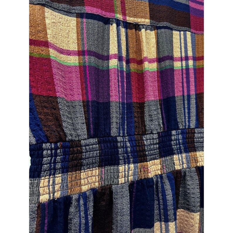 Pode incluir: Close-up de um tecido xadrez colorido. O padr&atilde;o apresenta uma mistura de cores, incluindo azul, rosa, castanho e bege. O tecido tem um detalhe de cintura el&aacute;stica franzida, adicionando textura e dimens&atilde;o ao design.