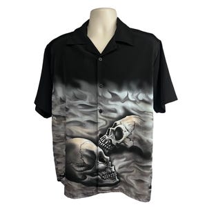Puede incluir: Camisa negra de manga corta con botones y estampado gráfico de dos calaveras sobre un fondo gris ahumado.