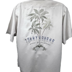 Pode incluir: Camisa bege clara de manga curta com um desenho de palmeiras nas costas. A camisa apresenta o texto "TOMMY BAHAMA" em azul marinho, juntamente com as palavras "WEEKENDS, WEEKDAYS, WHATEVER". O design inclui uma pequena ilustração de uma cena de praia.