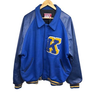 May include: Blue and navy blue varsity jacket with a yellow letter K on the front. The jacket has a leather sleeve and a ribbed collar, cuffs, and hem.