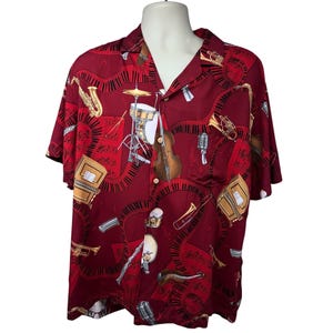 Puede incluir: Una camisa roja de manga corta con botones y estampado de instrumentos musicales. El estampado presenta una variedad de instrumentos, incluyendo una trompeta, un saxofón, un violín y tambores. La camisa tiene cuello y cierre de botones en la parte delantera.