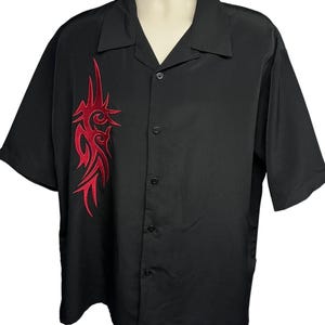 May include: Black short-sleeved shirt with a red tribal embroidery on the front. The shirt has a classic collar and button-down front. The shirt is designed for casual wear and has a relaxed fit.
