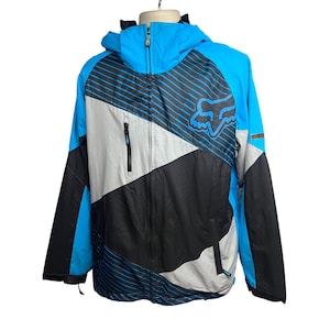 Puede incluir: Una chaqueta azul, negra y blanca con un logotipo de Fox Racing en la parte delantera. La chaqueta tiene una cremallera y una capucha.
