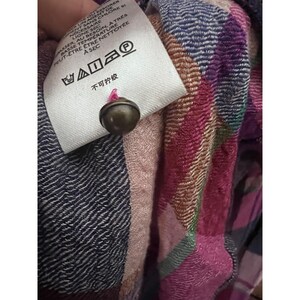Pode incluir: Close-up de um tecido xadrez multicolorido com uma etiqueta branca e um pequeno bot&atilde;o redondo de cor bronze. A etiqueta tem instru&ccedil;&otilde;es de cuidado em franc&ecirc;s e chin&ecirc;s. O tecido apresenta tons de rosa, roxo, azul e verde.