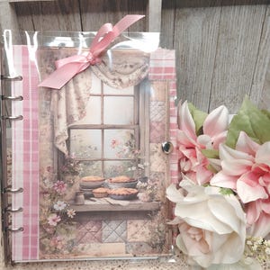 May include: A decorative scrapbook with a pink ribbon closure. The cover features a vintage illustration of a window with pies on a shelf. The scrapbook has a pink and white gingham pattern on the spine and edges, with a floral design.