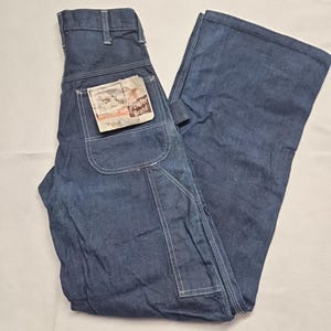 May include: Dark blue denim work pants with white stitching and a carpenter pocket. The pants have a tag that reads "Pointer Brand". The pants are folded and lying on a white surface.