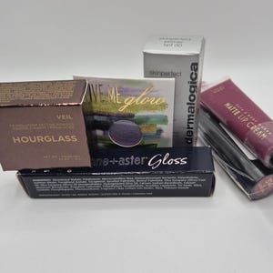 Op de afbeelding: Een verzameling schoonheidsproducten, waaronder Hourglass transparante setting powder, een skinperfect primer, een matte lip cream en oogschaduw. De producten zitten in verschillende containers en verpakkingen, met zichtbare tekstlabels.