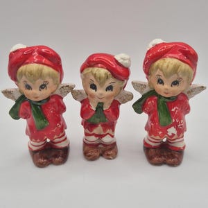 Peut inclure: Trois figurines en céramique de lutins de Noël. Chaque lutin porte une tenue rouge avec des bordures blanches, une écharpe verte et un chapeau rouge avec un pompon blanc. Les lutins ont les cheveux blonds, des visages peints et de petites ailes dorées.