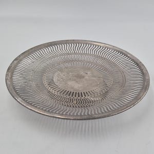 Puede incluir: Una bandeja redonda de metal plateado con un centro circular sólido rodeado por un borde decorativo ancho con un patrón repetitivo de recortes ondulados verticales finos que se extienden hasta el borde exterior liso y elevado.