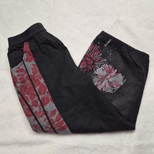 May include: Black capri trousers with an elasticated waistband. The trousers feature a grey and red floral design on the side and a large floral print on the leg. The fabric appears to be a soft denim.