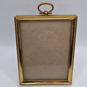 May include: A rectangular picture frame with a polished gold-tone metal finish. The frame features a simple, clean-lined border and a matching gold-tone oval hanging loop attached to the top center. The interior contains a plain, light beige backing.