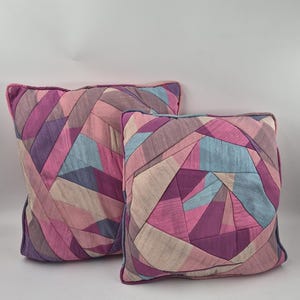 May include: Two square throw pillows featuring a patchwork design of fabric strips in shades of pink, purple, blue, and beige. The pillows have a visible piped edge and a textured, woven appearance with a geometric, multi-directional pattern.