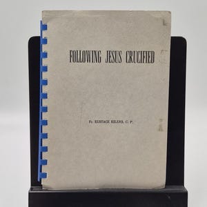 May include: A book titled "Following Jesus Crucified" by Fr. Eustace Eilers, C. P. The book has a light grey cover with black text and is bound with a blue plastic comb. It is displayed on a black stand.