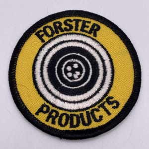 May include: A circular patch with a yellow background and a black border. The center features concentric black and white circles. The text "FORSTER" arches across the top, and "PRODUCTS" is at the bottom, both in black.
