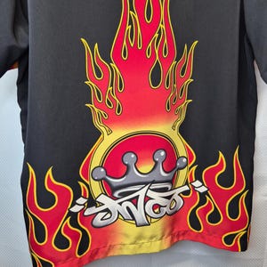 May include: Black short-sleeved shirt with a flame design in red, yellow, and orange. The design includes a crown and stylized text. The shirt's bottom edge has a yellow and orange gradient.