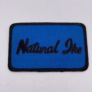 May include: A rectangular patch with a blue background and black stitched border. The words "Natural Ike" are embroidered in black cursive script across the center of the patch. The patch is likely designed to be sewn onto clothing or accessories.