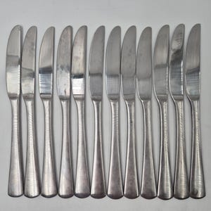 May include: A collection of twelve stainless steel butter knives. Each knife features a rounded blade and a long, tapered handle. The blades are slightly curved with a smooth, polished finish, suitable for spreading butter or other soft foods.