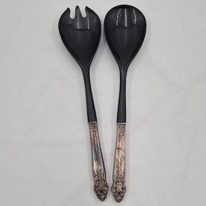 May include: A pair of salad serving utensils consisting of a fork and a spoon. Both pieces feature glossy black bowls and handles with ornate, metallic silver-toned floral detailing at the base. The design combines a modern dark finish with vintage-style metal accents.