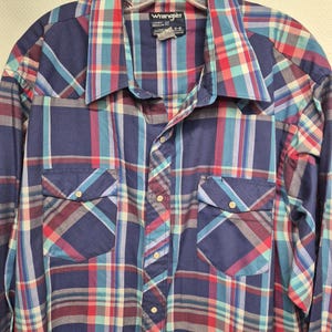 May include: A long-sleeved, button-down shirt with a plaid pattern in shades of navy blue, red, teal, and white. The shirt has two front pockets and a collar. The brand "Wrangler" is visible on a tag near the collar.