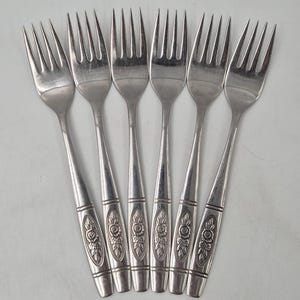 May include: A collection of seven silver forks. Each fork has a decorative floral pattern on the handle. The tines are long and thin. The forks are arranged in a fan shape against a white backdrop.