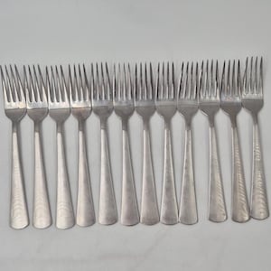 May include: A collection of silver forks arranged in a row. Each fork has four tines and a long, tapered handle. The forks are made of a shiny, metallic material, likely stainless steel, and are suitable for a formal dining setting.