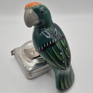 May include: A hand-painted wooden parrot figurine in shades of green, black, and orange, with a silver beak and a silver tape measure. The parrot has detailed feather patterns and is perched on top of the tape measure. The figurine is a decorative item.