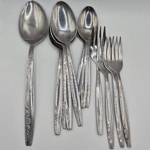 Peut inclure: Ensemble de couverts argentés vintage, comprenant des cuillères et des fourchettes, disposés sur une surface blanche. Les couverts présentent un motif floral sur les manches. Les cuillères ont une forme de bol ovale. Les fourchettes ont quatre dents.
