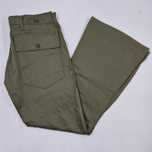 May include: Olive green trousers featuring a wide-leg silhouette, a structured waistband with belt loops, and a large rectangular patch pocket on the thigh secured with a single dark button. The fabric has a smooth, uniform texture throughout.