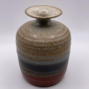 Może przedstawiać: Ręcznie robiony ceramiczny wazon o szerokim, okrągłym korpusie i rozkloszowanej górze. Wazon posiada poziome pasma kolorów, w tym beżowy, brązowy, niebieski, czarny i czerwony. Wazon ma około 18 cm wysokości.