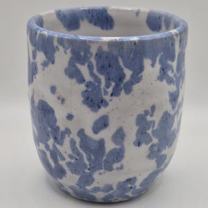 Peut inclure: Une tasse en céramique présentant une base blanche avec un motif irrégulier bleu tacheté et marbré sur toute la surface extérieure. Le récipient a une forme cylindrique avec une base légèrement arrondie et un bord ouvert et lisse.